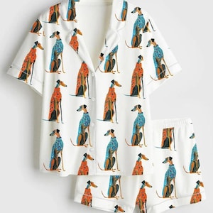 May include: A white pajama set consisting of a short-sleeved button-down shirt with a notched collar and matching shorts. The fabric features a repeating pattern of greyhound dogs wearing either orange polka-dot or blue patterned sweaters.