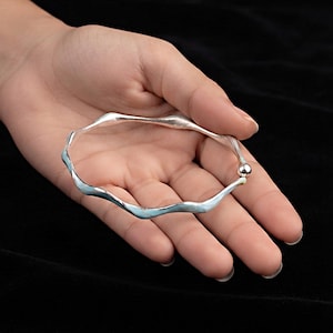 May include: A silver bangle bracelet with a wavy, undulating design. The bracelet features a smooth, polished metallic finish and a single small spherical bead detail at the point where the two ends of the wavy band meet to form a circular shape.