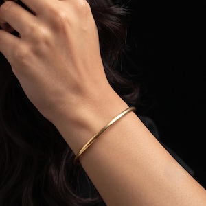 Op de afbeelding: Een eenvoudige, dunne, ronde armband van goudkleurig metaal met een gladde, matte afwerking. De armband is een massieve, doorlopende ring die om de pols zit, met een strak en onversierd ontwerp zonder zichtbare sluitingen, scharnieren of patronen.