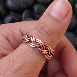 May include: A copper ring featuring a continuous braided or woven metal design. The band is constructed from multiple strands of polished copper wire intertwined to create a repeating pattern that encircles the entire circumference of the ring.