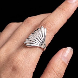 May include: A polished silver ring featuring a wide, fan-shaped design with multiple raised, vertical ridges that radiate outwards from the base of the band. The metallic surface reflects light, highlighting the curved, shell-like structure of the piece.