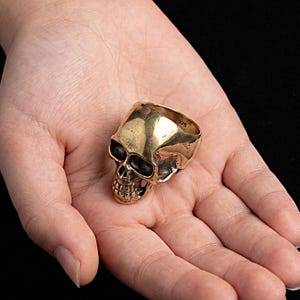 May include: A gold-toned metal ring shaped like a human skull. The ring features a polished, smooth cranium surface with darkened, recessed details around the eye sockets, nasal cavity, and teeth. The interior band is clearly stamped with the number 375.