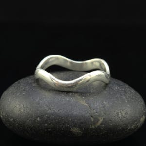 Puede incluir: Un anillo de tono plateado pulido que presenta un diseño de banda continua, suave y ondulada. La superficie metálica es reflectante y de color uniforme, mostrando una forma fluida y curva que rodea el centro de la banda del anillo.