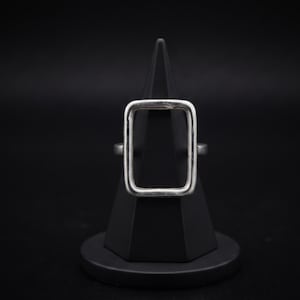 Op de afbeelding: Een zilverkleurige ring met een opvallend, verticaal georiënteerd rechthoekig frameontwerp. De band is glad en metaalachtig en toont een strakke, in het midden open geometrische vorm die op de displaystandaard is geplaatst.