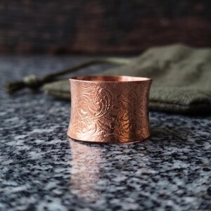 Könnte beinhalten: Ein breiter Kupferring mit einem detaillierten, geprägten Blumenmuster auf der gesamten Außenfläche. Das Metall hat eine warme, rotbraune metallische Oberfläche mit einer leicht konkaven Form, die komplizierte Texturen und Glanz zeigt.