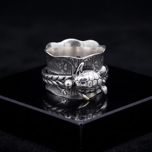 May include: A silver spinner ring featuring a central band with a raised sea turtle motif and a braided texture. The wider base band displays an engraved floral pattern with vertical lines, finished with a scalloped edge along the top rim.