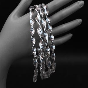 Könnte beinhalten: Ein Set aus drei silberfarbenen Armreifen mit einem durchgehenden, gedrehten Spiraldesign. Jeder Armreif hat eine polierte, reflektierende Metalloberfläche, die das Licht einfängt und eine Reihe sich wiederholender Kurven erzeugt.