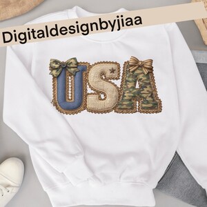May include: White sweatshirt featuring the word "USA" in a decorative design. The letters are embellished with bows and patterns, including denim, burlap, and camouflage. The text "Digitaldesignbyjiaa" is printed across the top.