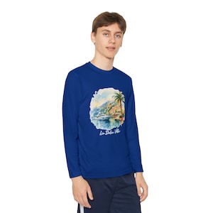 Pode incluir: Uma camisa azul de manga comprida com um desenho em aquarela de uma cena costeira com um barco, uma palmeira e edifícios. O texto "La Dolce Vita" está impresso abaixo. A camisa é de cor azul lisa.
