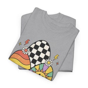 May include: Two gray sweatshirts are folded and stacked. The top shirt features a colorful graphic design with a checkered oval, rainbow stripes, a smiling sun, and daisies. The design also includes lightning bolts and stars.