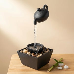 May include: A black ceramic teapot fountain with water flowing into a small bowl set in a square, black planter filled with pebbles. The fountain sits on a wooden surface, with bamboo and stones nearby.