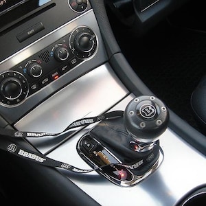 Op de afbeelding: Interieurdetail van een auto met een zwarte versnellingspookknop met het "B"-logo, een zilveren console en een zwart koord met "BRABUS"-tekst. Het dashboard bevat klimaatregelingsknoppen en -knoppen.