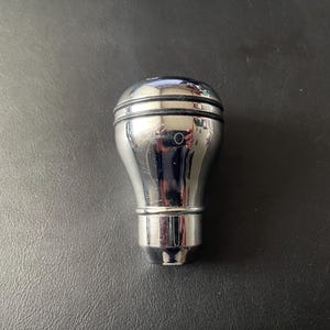 May include: A polished chrome gear shift knob, featuring a rounded top and a cylindrical base. The reflective surface mirrors the surroundings. Designed for automotive applications.