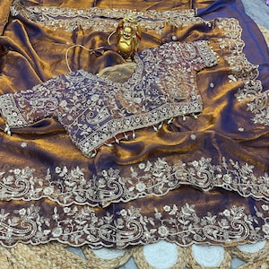 May include: A luxurious, shimmering brown saree with intricate gold embroidery. The saree features a matching blouse with detailed floral patterns and delicate pearl accents. The fabric has a rich, flowing texture, perfect for special occasions.