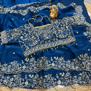 May include: A deep blue saree with intricate gold embroidery. The saree features a matching blouse with detailed floral and paisley patterns. A small gold Buddha statue and a gold necklace are also visible.