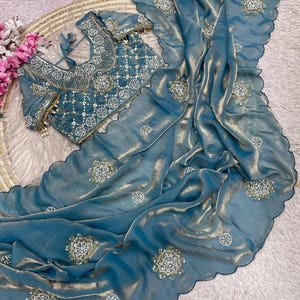 Peut inclure: Un ensemble sari de couleur sarcelle avec des broderies complexes. L'ensemble comprend un chemisier et un sari drapé, tous deux avec des motifs et des embellissements détaillés. Le sari a un bord festonné et est orné de motifs floraux. Le chemisier a un design à nouer dans le dos.