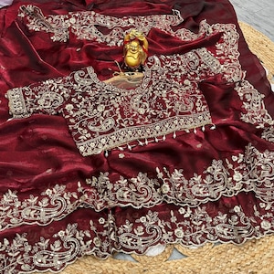 Könnte beinhalten: Ein bordeauxfarbener Sari mit aufwendiger Goldstickerei. Der Sari weist detaillierte Blumen- und Paisley-Muster an den Rändern und auf der Bluse auf. Eine kleine goldene Statue befindet sich auf dem Sari.