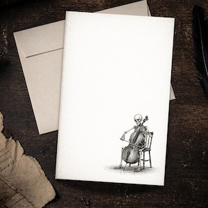 May include: A blank white card with a black and white illustration of a skeleton playing a cello. The skeleton is seated on a chair. A beige envelope is behind the card. A feather is on the right side of the image.