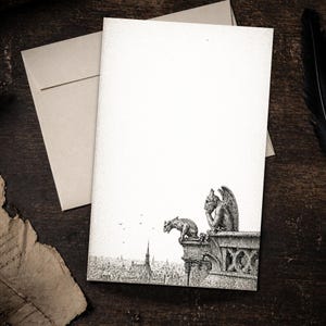 May include: Blank stationery set with a black and white illustration of gargoyles on a building overlooking a city. Includes a matching envelope, set against a dark wood background.