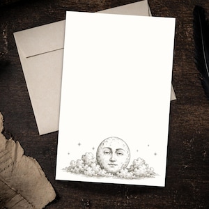 May include: A blank stationery card with a detailed illustration of a full moon with a face, emerging from clouds. The card is accompanied by a matching envelope. The design is in black and white, with a vintage aesthetic.