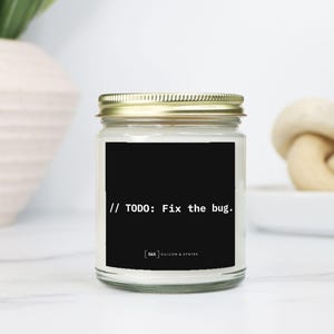 Może przedstawiać: Świeca w szklanym słoiku ze złotą pokrywką i czarną etykietą. Na etykiecie widnieje napis "// TODO: Fix the bug." białym tekstem. To element wystroju wnętrz.