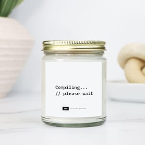 May include: A clear glass jar candle with a gold lid. The candle has a white label with the text "Compiling... // please wait" in black. The bottom of the label has the logo for "Silicon & Syntax".