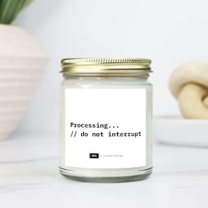May include: A glass jar candle with a gold lid. The label displays "Processing... // do not interrupt" in black text. The candle sits on a white surface. A white vase and a decorative knot are in the background.