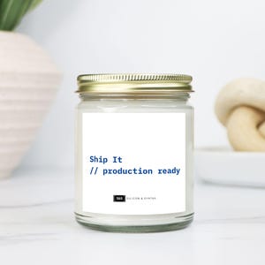 Op de afbeelding: Een glazen potkaars met een gouden deksel. Het etiket leest "Ship It // production ready" in blauwe tekst. De kaars staat op een wit oppervlak, met een witte vaas en een decoratieve knoop op de achtergrond.