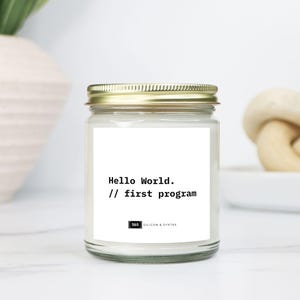 Puede incluir: Una vela en tarro de cristal transparente con tapa dorada. La etiqueta dice "Hello World. // first program" en texto negro. La vela está sobre una superficie blanca, con un jarrón blanco y un nudo decorativo en el fondo.