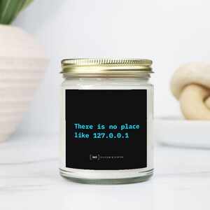 May include: A glass jar candle with a gold lid. The label reads "There is no place like 127.0.0.1" in teal on a black background. A home decor item.