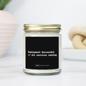 Könnte beinhalten: Ein Glas-Kerzenglas mit goldenem Deckel. Die Kerze hat ein schwarzes Etikett mit weißem Text, der lautet: "Deployment Successful // all services running". Auf der Unterseite des Etiketts steht der Text "S&S SILICON & SYNTAX".