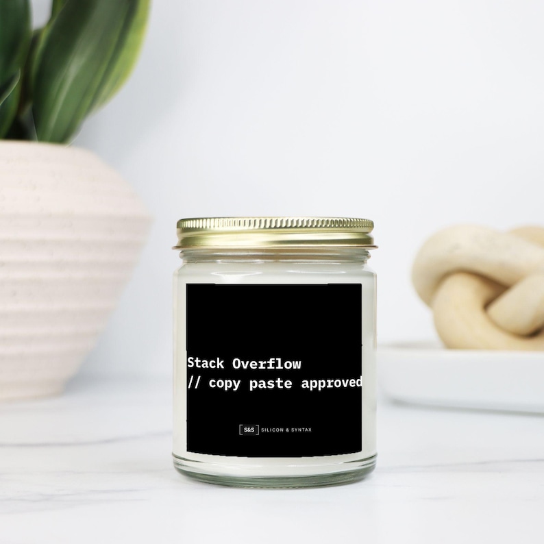 May include: A clear glass jar candle with a gold lid. The candle has a black label with the text "Stack Overflow // copy paste approved". The candle is on a white surface.