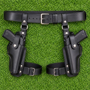 May include: A black leather double holster rig featuring a waist belt with a silver-toned metal buckle and two drop-leg holsters. Each holster is secured with a snap button strap and includes an adjustable leg strap with a silver-toned metal buckle.