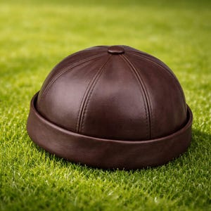 Puede incluir: Gorra de cuero marrón con corona redondeada y ala corta y plana. La gorra tiene costuras verticales y un pequeño botón en la parte superior. Se encuentra sobre hierba verde.