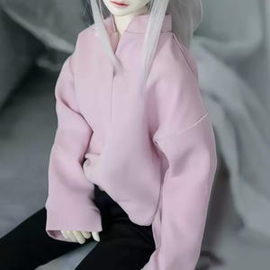 May include: A pale pink long-sleeved top with a wide collar and a relaxed fit. The top is worn with black trousers. The image shows a doll wearing the outfit. The doll has white hair.