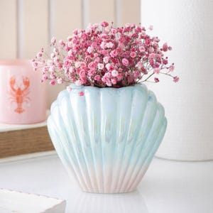 Może przedstawiać: Wazon w kształcie muszli w odcieniach błękitu i różu, wypełniony różowymi kwiatami. Wazon ma perłowe wykończenie. W tle różowy świecznik z motywem homara.