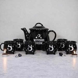Puede incluir: Un juego de té de cerámica negra con una tetera y seis tazas. La tetera tiene las palabras "Witches Brew" en blanco. Las tazas y la tetera tienen un diseño de luna creciente y estrellas blancas. Cada taza tiene el número "5" en blanco.