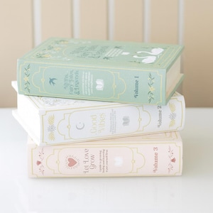 Puede incluir: Tres libros decorativos apilados. El libro superior es de color verde claro con el texto "Myths, Fairy Tales & Legends" y "Volume 1". El libro del medio es blanco con "Good Vibes" y "Volume 2". El libro inferior es rosa con "Let Love Grow" y "Volume 3".