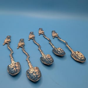 Könnte beinhalten: Fünf silberfarbene Löffel mit einem von der Natur inspirierten Design. Jeder Löffel hat eine blattförmige Schale und einen astähnlichen Griff, der von einem Vogel gekrönt wird. Die Löffel sind auf blauem Hintergrund angeordnet.