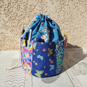 Può includere: Un portapenne e matite con coulisse e vari pannelli di tessuto colorati. I pannelli presentano motivi geometrici, floreali e a farfalla nei toni del blu, rosa e verde. Penne e matite sono visibili.