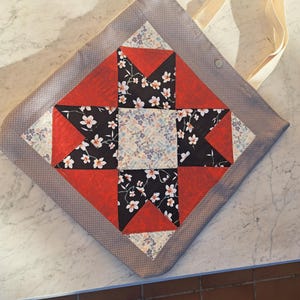 Peut inclure: Un sac fourre-tout fait main avec un motif matelassé. Le sac présente un motif carré central en tissus rouge, noir floral et floral clair. Le bord extérieur est gris à pois blancs et possède des anses crème.