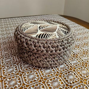 Può includere: Un cestino rotondo all'uncinetto nei toni del marrone, con un coperchio in legno con un motivo a foglie. Il cestino è appoggiato su una tovaglia di pizzo bianco, creando un contrasto di trame e colori. Adatto per riporre oggetti o decorare.