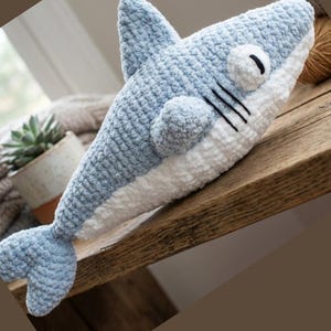 May include: A handmade, plush shark toy in shades of light blue and white. The shark features a textured, crocheted body, a dorsal fin, and a tail fin. It has black stitched details for the eyes and gills, and a round belly.
