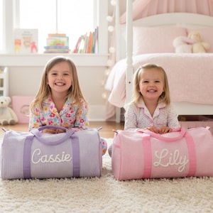 May include: Two cylindrical duffel bags made of striped fabric. The left bag is light purple with white stripes and the name Cassie embroidered in white. The right bag is light pink with white stripes and the name Lily embroidered in white.
