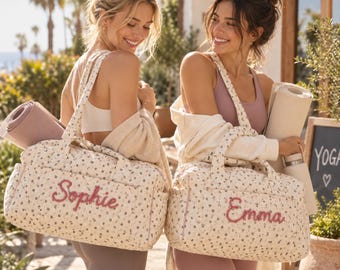 May include: Two quilted duffel bags featuring a small floral pattern on a cream background. Each bag has two top handles and is personalized with embroidered names in pink script: one reads Sophie and the other reads Emma.