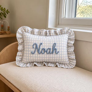 May include: A rectangular throw cushion featuring a light blue and white gingham check pattern with a ruffled border. The name Noah is embroidered in a raised, textured blue script font across the centre of the cushion surface.