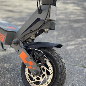 Op de afbeelding: Een close-up van een zwarte en grijze elektrische scooter. De scooter heeft een oranje accent op de voorvork en een zwarte band met een schijfrem. Ontworpen voor gebruik buitenshuis.