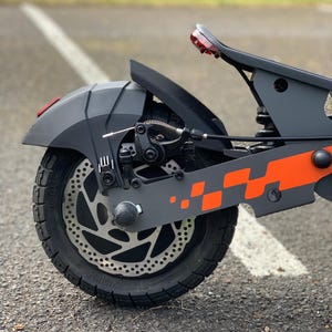Op de afbeelding: Close-up van een grijze en zwarte elektrische scooter. De scooter heeft een oranje geblokt ontwerp, een schijfrem en een zwarte band. Een rode reflector is zichtbaar aan de achterkant. De scooter staat op een asfalt oppervlak.