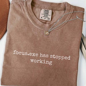May include: A brown short-sleeved t-shirt with a crew neckline and the white text focus.exe has stopped working printed on the front. The shirt features a Comfort Colors brand label at the inner neck and is paired with a thin silver-toned necklace.