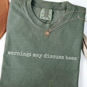 May include: A moss green short-sleeve t-shirt with a crew neckline and the white text "warning: may discuss bees" printed across the chest. The shirt features a Comfort Colors brand label inside the collar and a thin gold-tone chain necklace.